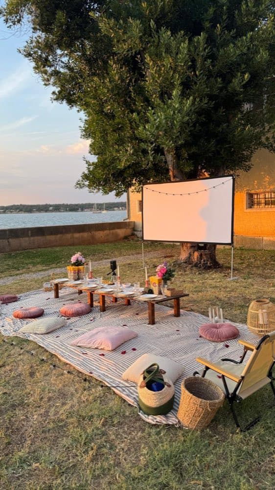 Niedrige Tafel und Kissen auf gestreifter Decke vor einer Leinwand unter einem Baum am Meer im Abendlicht