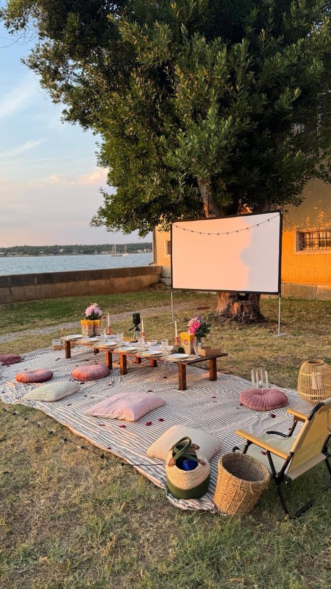 Long low table and cushions on a striped blanket facing a projector screen under a tree by the sea at dusk