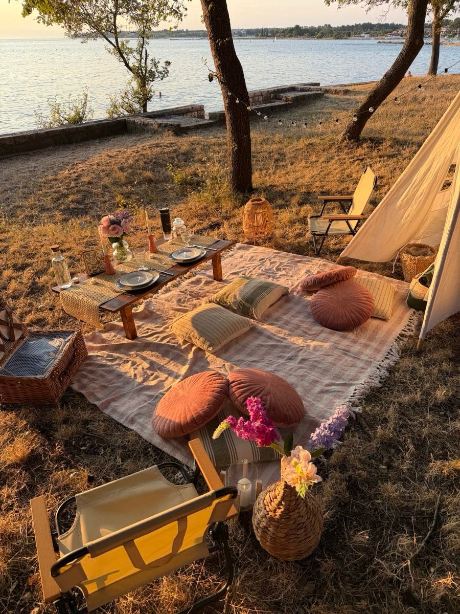 Low table set for two beside a teepee, with round cushions, a picnic hamper and the sea in the evening sun