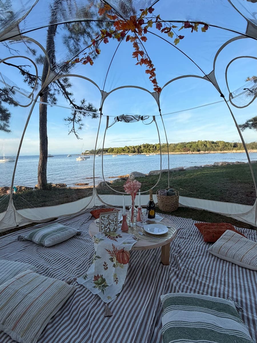Inside a clear igloo dome: low round table with candles and wine, autumn leaf garland, sea with boats beyond