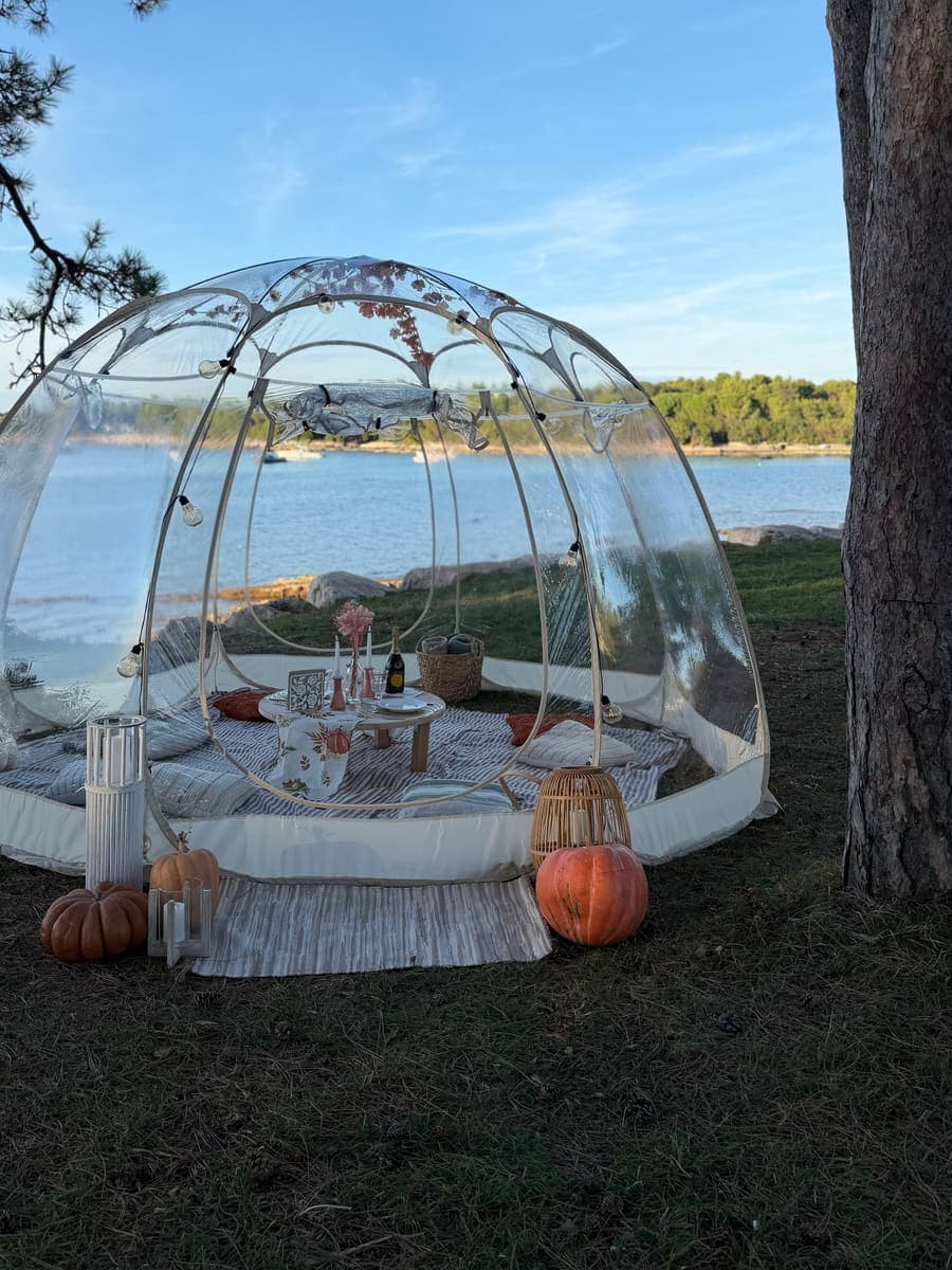 Transparente Iglu-Kuppel auf einer Wiese am Meer zwischen Kiefern, mit Kürbissen und Laternen am Eingang
