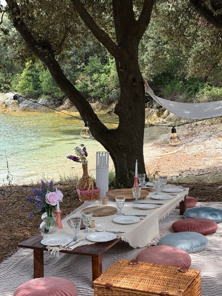 Lange niedrige Tafel mit Tellern und Gläsern unter einem Baum an einer Bucht, mit Hängematte und Lichterkette