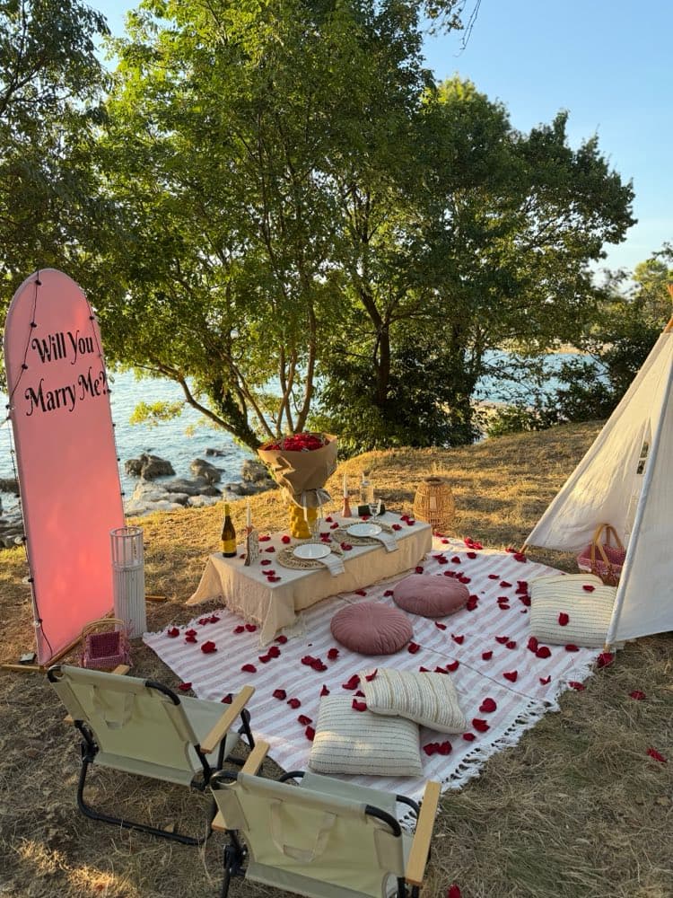Pink “Will You Marry Me?” sign beside a low table for two with red roses, rose petals and a teepee by the sea